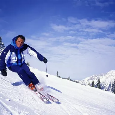 Ein Skifahrer fährt mit Schwung die verschneite Piste hinunter. Im Hintergrund sind schneebedeckte Berge und ein klarer Himmel zu sehen.