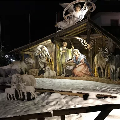 Eine festliche Krippe mit Figuren von Maria, Josef und dem Baby Jesus steht in einem verschneiten Umfeld. Im Hintergrund leuchtet ein Engel und die gesamte Szene wirkt weihnachtlich und besinnlich.