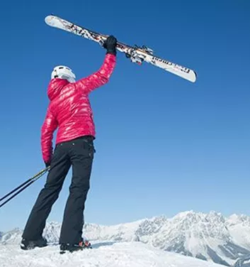 A person in a red ski jacket is lifting a pair of skis high. In the background, snow-covered mountains can be seen under a clear blue sky.