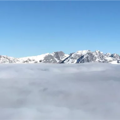 Eine beeindruckende Berglandschaft erhebt sich über einer dichten Wolkenschicht. Der Himmel ist klar und blau.