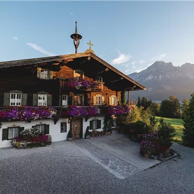 Ein traditionelles bayerisches Haus mit Blumenkästen und einem schönen Garten. Im Hintergrund sind majestätische Berge und ein klarer Himmel zu sehen.