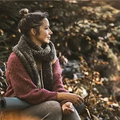 Eine Frau sitzt in einem herbstlichen Wald und betrachtet die Umgebung. Sie trägt einen warmen Pullover und einen Schal.