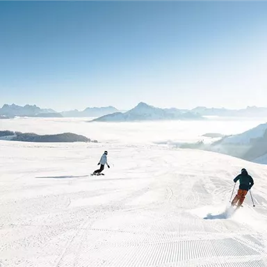 Drei Skifahrer fahren durch eine schneebedeckte Landschaft. Im Hintergrund sind majestätische Berge und ein klarer blauer Himmel zu sehen.