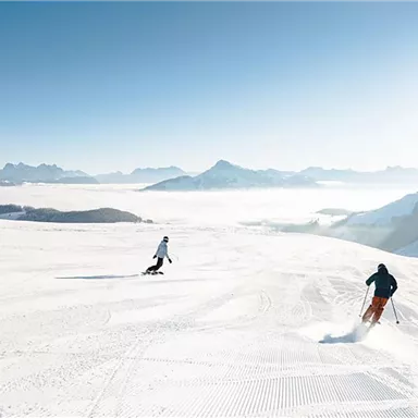 Three skiers enjoy a snow slope under a clear sky. Majestic mountains can be seen in the background.