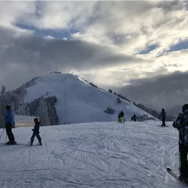 Eine schneebedeckte Skipiste mit mehreren Skifahrern. Im Hintergrund ist ein hügeliger Berg und ein bewölkter Himmel zu sehen.