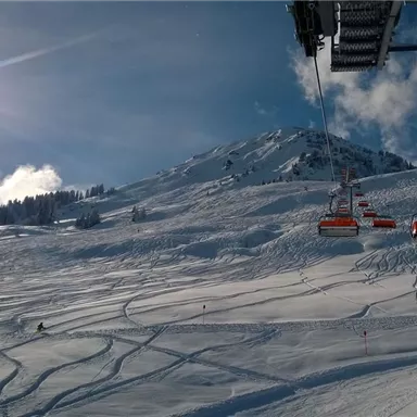 Ein schneebedeckter Hang mit Skizügen und einer sonnigen, klaren Himmel. Im Hintergrund ist ein Skilift zu sehen.