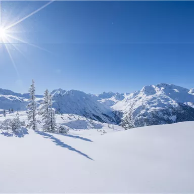 A snow-covered landscape with majestic mountains and a clear blue sky. The sun shines brightly over the winter scenery.