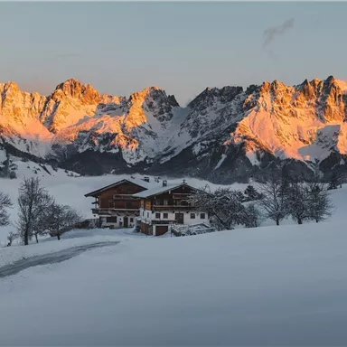 A picturesque winter landscape with snow-covered meadows and majestic mountains. A traditional chalet stands in front of the impressive peaks illuminated by the evening sun.