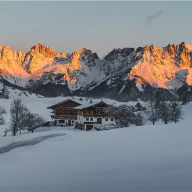 A picturesque winter landscape with snow-covered mountains and a traditional wooden house. The sun rays illuminate the mountain peaks in orange.