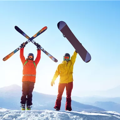Two skiers are standing on a snowy peak, holding their skis and a snowboard up in the air. The sky is clear and blue, and the mountain landscape is visible in the background.