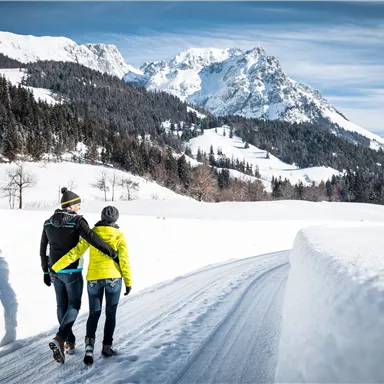 Ein Paar spaziert auf einem verschneiten Weg in den Bergen. Die Umgebung ist idyllisch mit schneebedeckten Bäumen und beeindruckenden Bergen im Hintergrund.