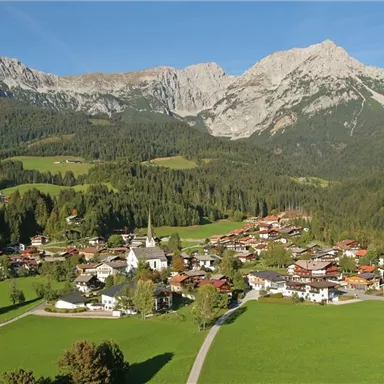 Eine malerische Berglandschaft mit einem kleinen Dorf inmitten grüner Wiesen. Im Hintergrund ragen hohe Berge unter einem klaren blauen Himmel empor.