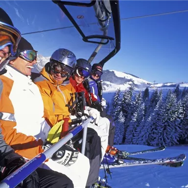 Five skiers are sitting on a chairlift in a snowy mountain landscape. In the background, there are pine trees and a clear blue sky.