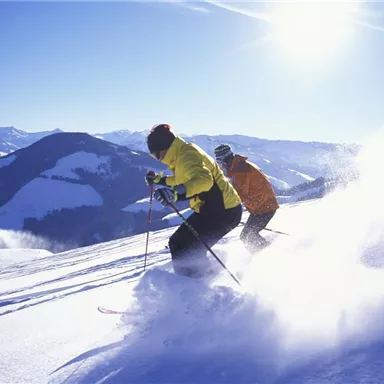 Zwei Skifahrer fahren die verschneite Piste hinunter. Die Landschaft ist von Bergen und klarem, sonnigem Himmel umgeben.