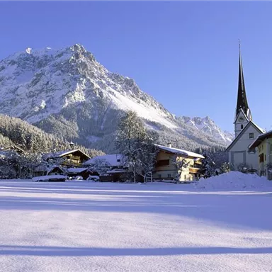 Eine winterliche Landschaft mit schneebedeckten Bergen und einem kleinen, charmanten Dorf. Im Vordergrund steht eine Kirche mit einem hohen Turm.