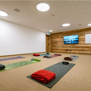 Ein heller, ruhiger Raum mit Yoga-Matten in verschiedenen Farben. An der Wand befindet sich ein Fernseher und die Atmosphäre ist entspannend.