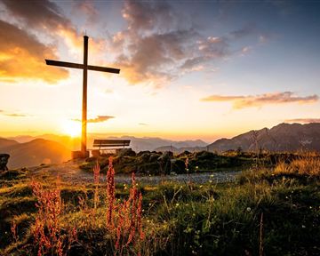 A large cross stands on a hill at sunset. The surroundings are beautiful with mountains and meadows visible.