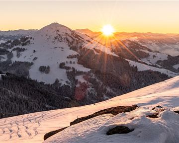 Eine verschneite Berglandschaft bei Sonnenaufgang. Die Sonne strahlt über die Berge und den Winterwald.