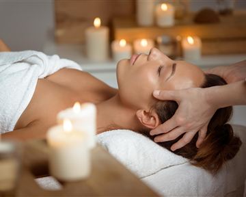 Eine entspannende Wellnessanwendung mit einer Frau, die eine Kopfmassege erhält. In der Umgebung sind Kerzen und ein ruhiges Ambiente zu sehen.