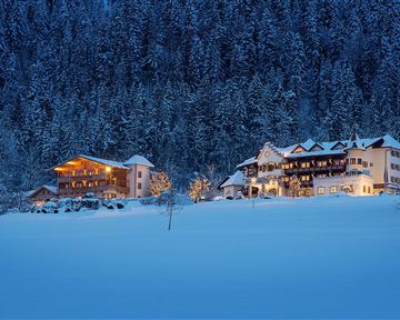 Eine winterliche Landschaft mit schneebedeckten Hügeln. Gemütliche Häuser leuchten sanft im Dämmerlicht.