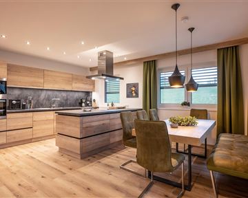 A modern kitchen with wooden shelves and a big dining table. The open atmosphere is emphasized by the elegant lamps and the green curtains.