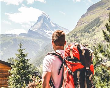 Ein Wanderer mit einem großen Rucksack blickt auf die beeindruckenden Berge. Die Landschaft ist grün und die Wolken ziehen am Himmel vorbei.