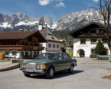 An old Mercedes-Benz drives through a picturesque village with traditional architecture. In the background, majestic mountains rise under a clear sky.