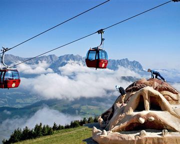An impressive mountain landscape with gondolas floating above the clouds. In the foreground stands a large sculpture of a face.