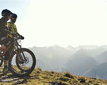 Two cyclists are standing on a high trail, admiring the mountain landscape. The sun is shining over the peaks, providing a breathtaking view.