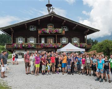 Eine große Gruppe von Menschen steht vor einem traditionellen, blumengeschmückten Chalet in den Bergen. Im Hintergrund sind grüne Hügel und ein bewölkter Himmel zu sehen.