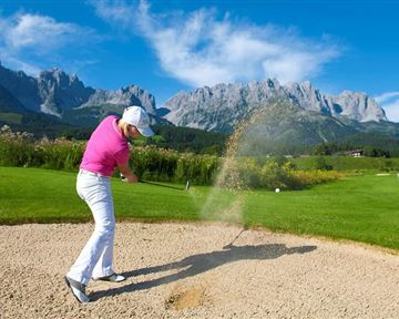 A golfer is hitting from a bunker on a beautiful golf course with impressive mountains in the background. The sky is clear and blue, the landscape is green and inviting.
