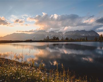 Ein malerischer See, umgeben von Bergen und blühenden Wiesen. Die untergehende Sonne spiegelt sich ruhig im Wasser.