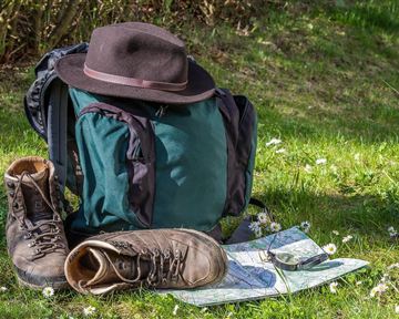 Ein Rucksack steht auf einer Wiese, neben einem Paar Wanderschuhe und einer Karte. Ein Hut liegt auf dem Rucksack und im Hintergrund sind frisches Gras und kleine Blumen zu sehen.