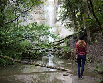 Eine Frau steht an einem kleinen Wasserfall in einem bewaldeten Gebiet. Der Wald ist grün und üppig, mit einem klaren Wasserbecken im Vordergrund.