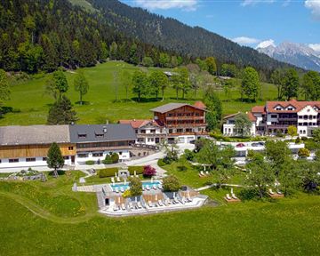 Eine malerische Landschaft mit einem Hotelkomplex, umgeben von Wäldern und Bergen. Im Vordergrund sind ein Poolbereich und grüne Wiesen zu sehen.