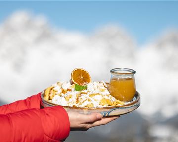 Ein Teller mit frischen, süßen Speisen, garniert mit einer getrockneten Orange und einem Glas Saft. Im Hintergrund sind schneebedeckte Berge sichtbar.