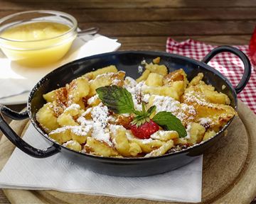 Ein köstlicher Kaiserschmarrn, serviert in einer Pfanne, mit Puderzucker, frischen Beeren und Minze. Dazu gibt es eine Schüssel Apfelmus.