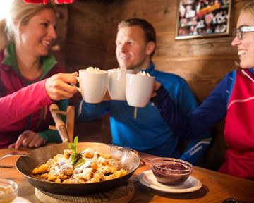 Three people clink glasses and enjoy a meal in a cozy cabin. On the table, there is a bowl with a delicious dessert.