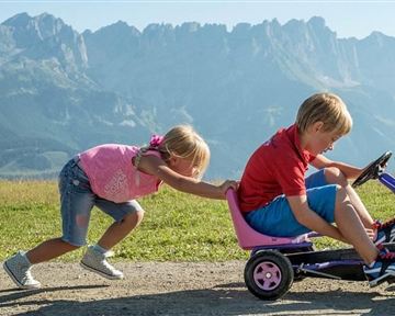 Ein Junge fährt auf einem kleinen Fahrzeug, während ein Mädchen ihn von hinten schiebt. Die Szene spielt vor einer beeindruckenden Bergkulisse.