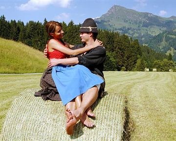 EinPaar sitzt auf einem Heuballen in einer grünen Wiese mit Bergen im Hintergrund. Sie lächeln sich an und genießen die schöne Landschaft.