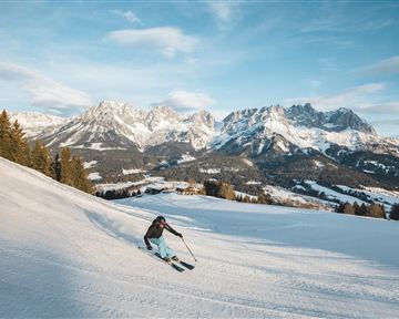 Ein Skifahrer fährt die schneebedeckte Piste hinunter. Im Hintergrund sind majestätische Berge und ein blauer Himmel zu sehen.