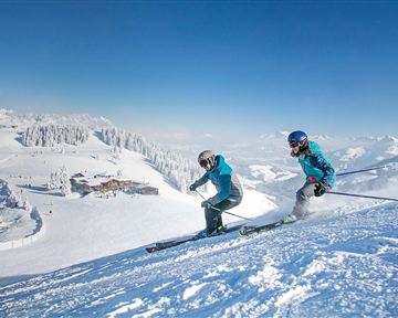 Two skiers are skiing down the snowy slope. In the background, there are snow-covered mountains and a blue sky.