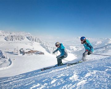 Zwei Skifahrer fahren die verschneite Piste hinunter. Im Hintergrund erstrecken sich schneebedeckte Berge und ein klarer blauer Himmel.