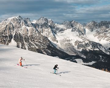 Two skiers are going down the snowy slope. In the background, impressive mountains can be seen under a gray sky.