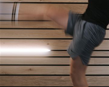 Ein Mann führt einen dynamischen Kick aus. Im Hintergrund sind holzverkleidete Wände und eine moderne Beleuchtung zu sehen.