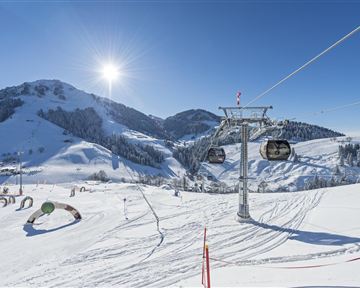 An idyllic winter landscape with skiing slopes and a chairlift. The sky is clear and the sun is shining over the snow-covered mountains.