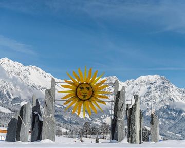 Eine winterliche Landschaft mit schneebedeckten Bergen und einem bunten Sonnensymbol in der Mitte. Rund um das Symbol stehen große Steine, die eine mystische Atmosphäre schaffen.