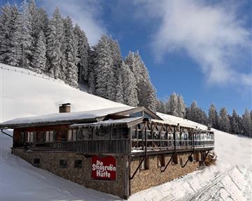 Eine gemütliche Berghütte im Schnee, umgeben von schneebedeckten Bäumen. Der Himmel ist klar und blau.