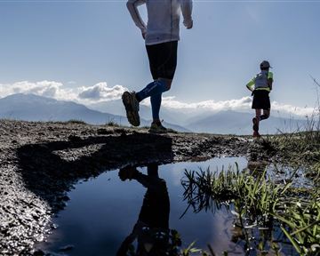 Two runners are jogging on an uneven path with a view of the mountains. In the foreground, the sky is reflected in a puddle.