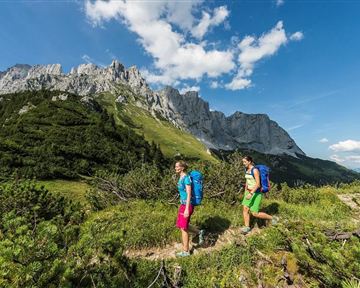 Zwei Wanderer gehen auf einem Pfad in den Bergen. Die Landschaft ist grün mit felsigen Gipfeln und klarem Himmel.
